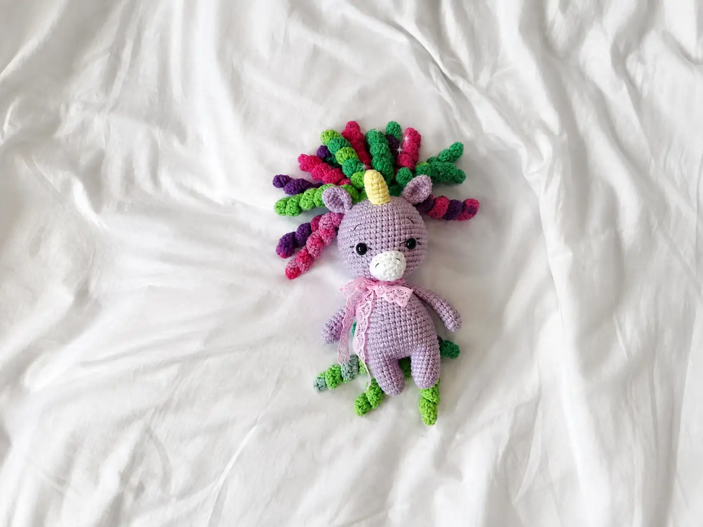 A purple crochet unicorn amigurumi with a pastel curled mane and pink ribbon, sitting in a woven basket.