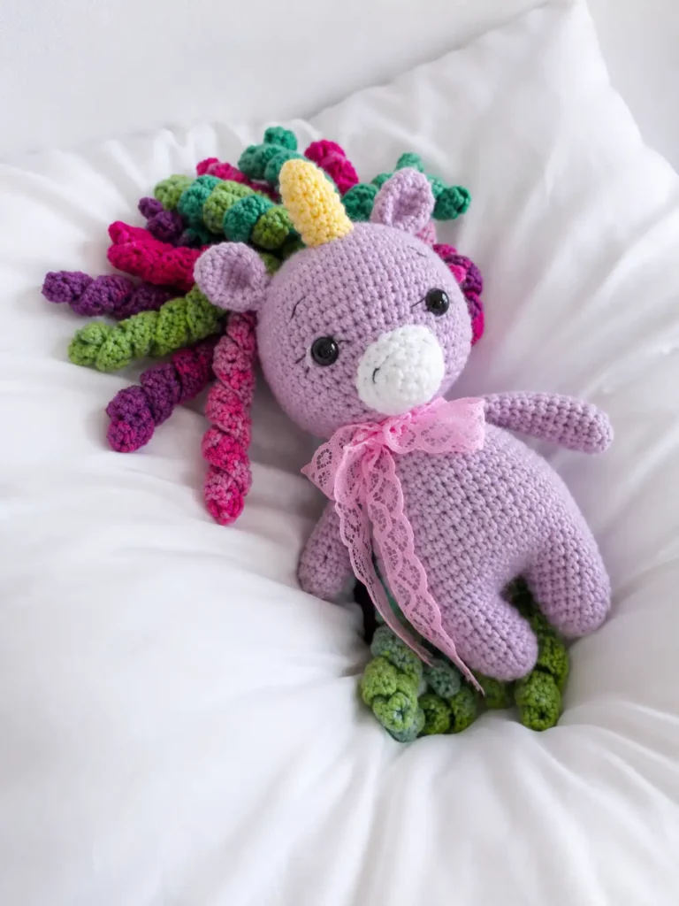 Close up of a cute unicorn amigurumi free pattern project in purple yarn with colorful curls, lying on a white pillow.