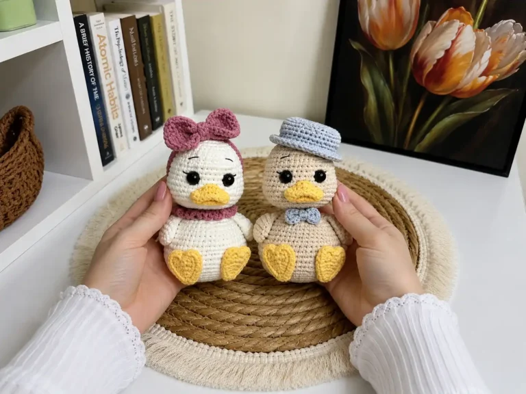 Two hands holding crochet duckling amigurumi dolls, one with a pink bow and one with a blue hat, outdoors.