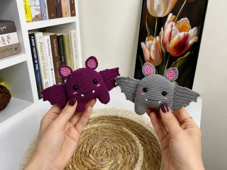 Hands holding purple and grey plush crochet bats made with fine yarn against a warm blurred room background.
