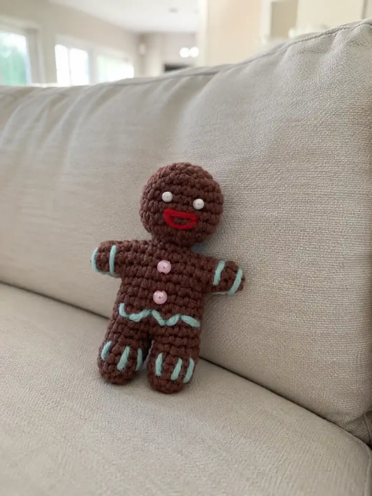 Crocheted brown gingerbread man amigurumi free pattern with pink beads resting against a textured beige couch.