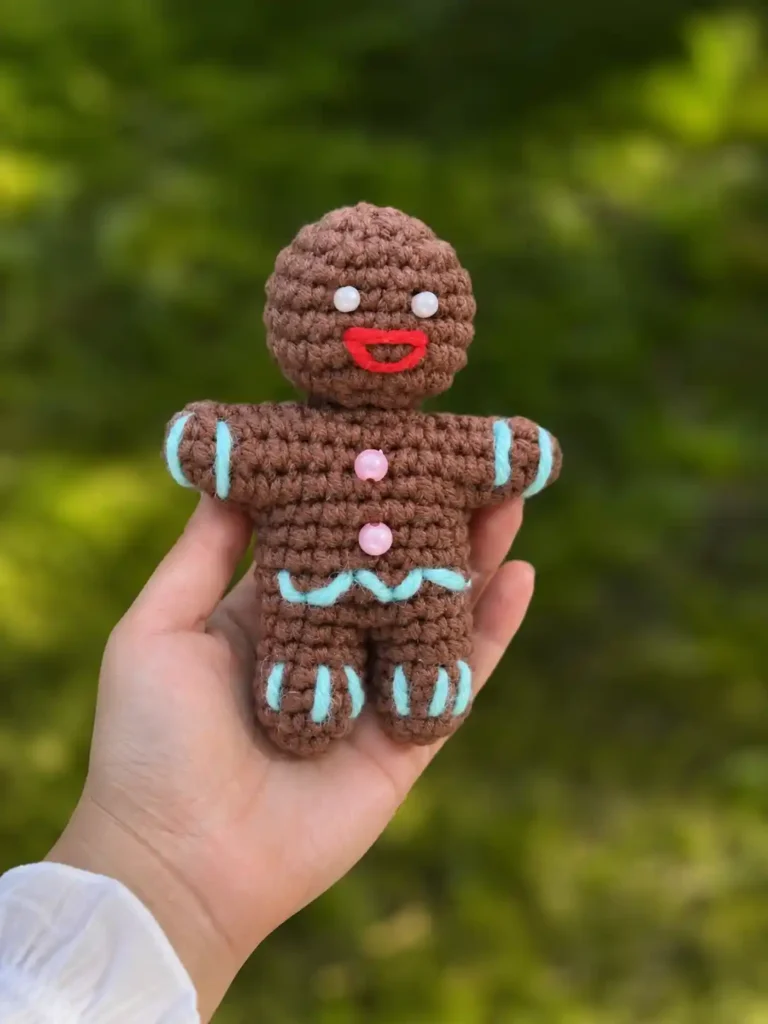 Hand holding a crocheted brown gingerbread man amigurumi free pattern outdoors against a blurred green background.
