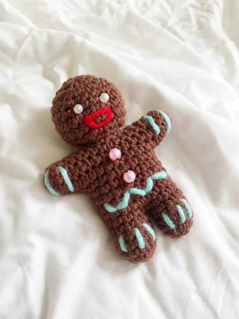 Crocheted brown gingerbread man amigurumi free pattern lying flat on a softly wrinkled white fabric bedsheet.