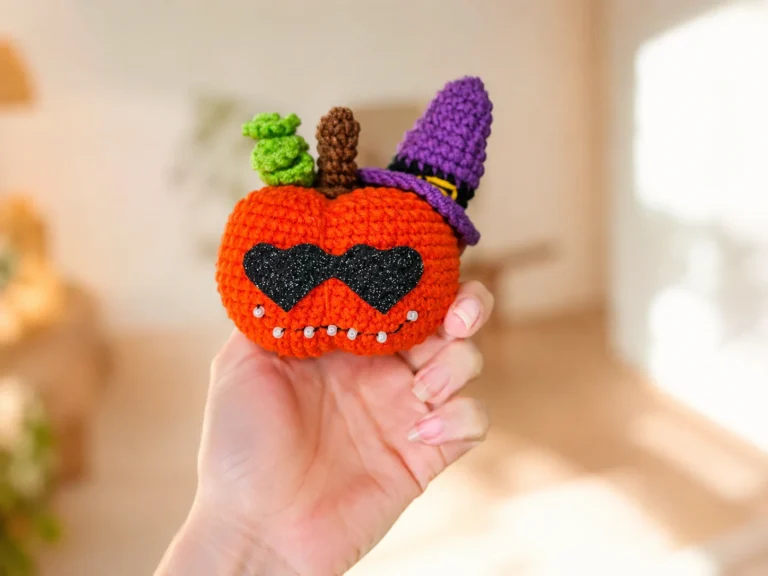 Orange crochet pumpkin amigurumi with heart-shaped black sunglasses and a purple witch hat on a wood table.
