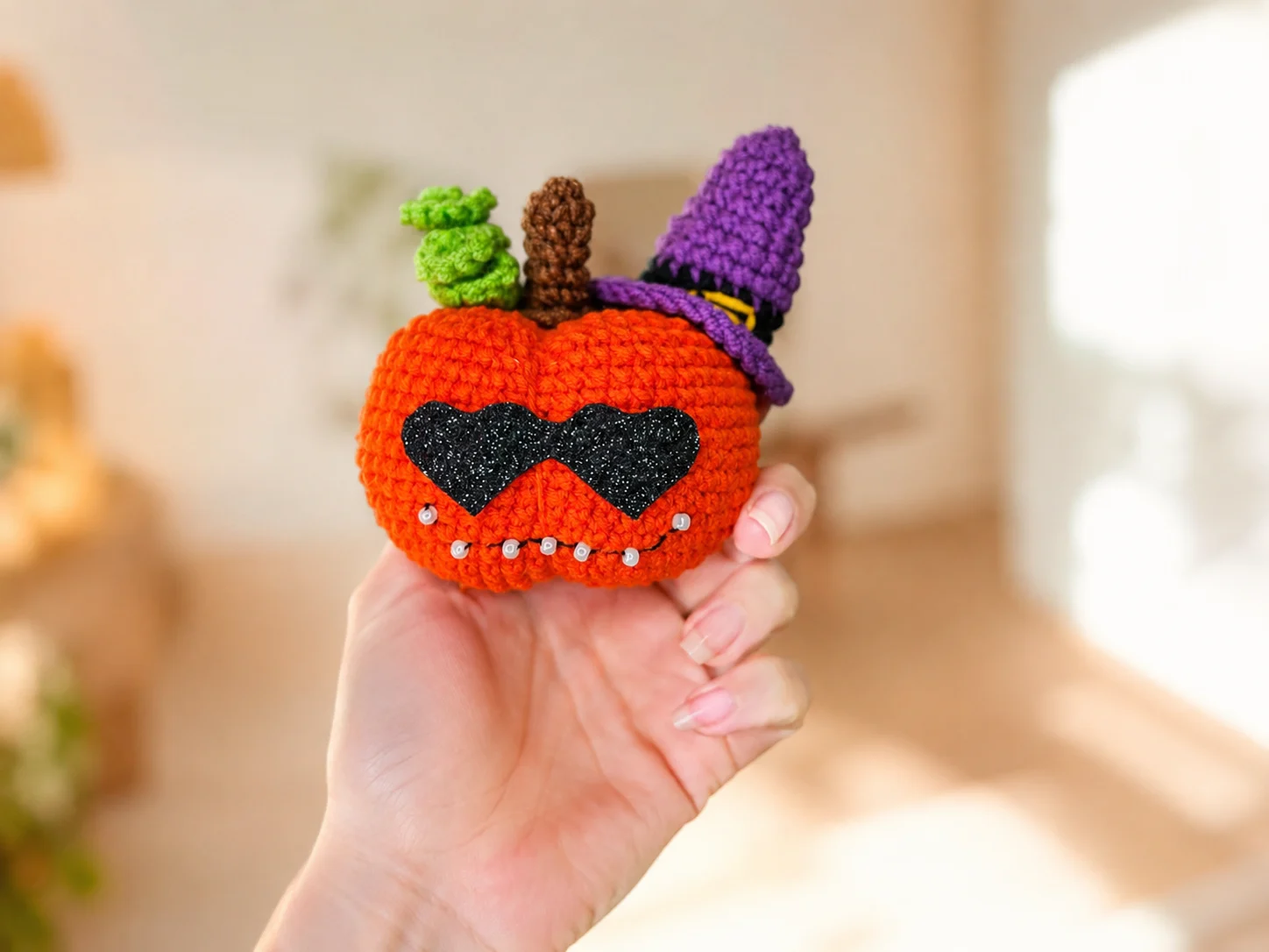 Orange crochet pumpkin amigurumi with heart-shaped black sunglasses and a purple witch hat on a wood table.