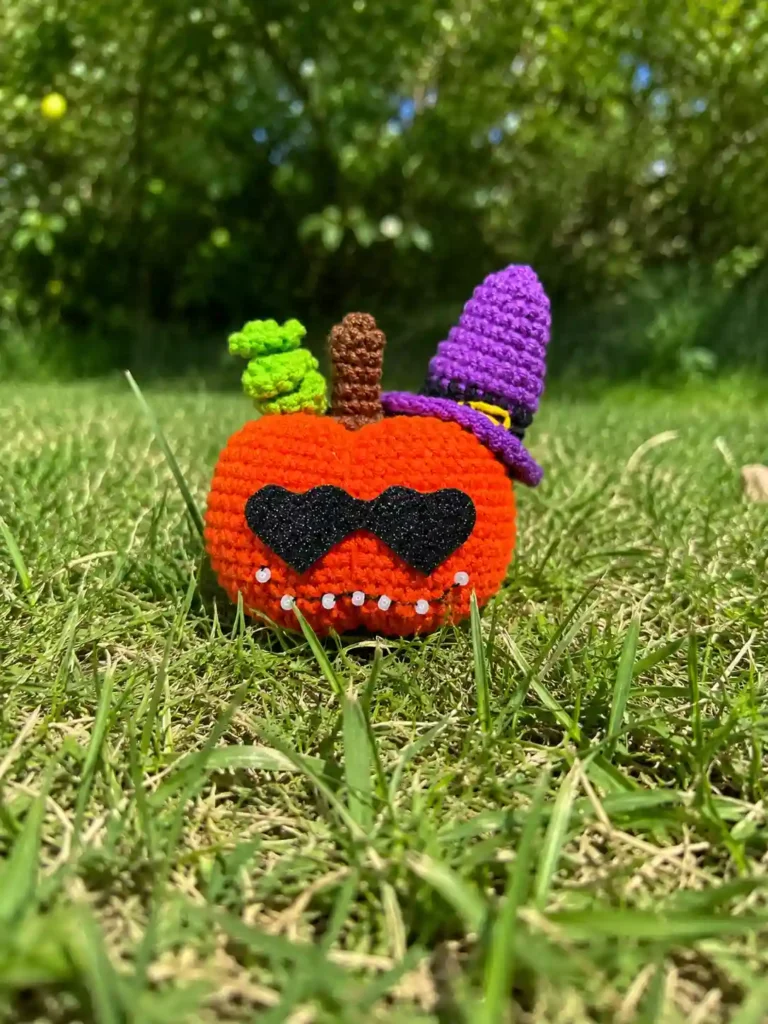 The glam pumpkin amigurumi free pattern outdoors. Orange yarn toy with a purple witch hat and black glasses in green grass.