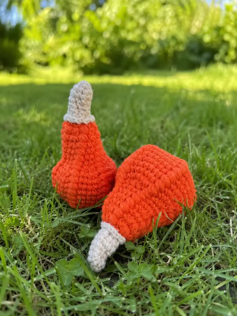 Two orange crocheted gourds with off-white stems from a halloween pumpkin crochet pattern rest in bright green grass.