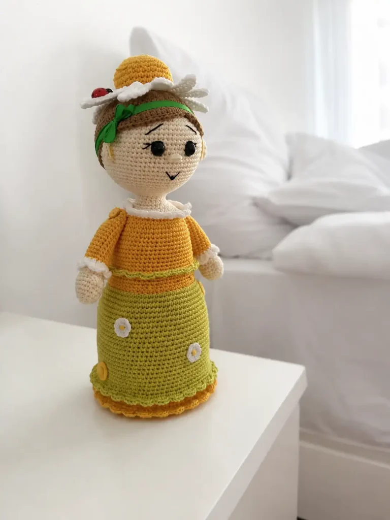 A yellow and green cotton yarn doll sits on a white desk by a bed, modeling the chamomile doll amigurumi free pattern.