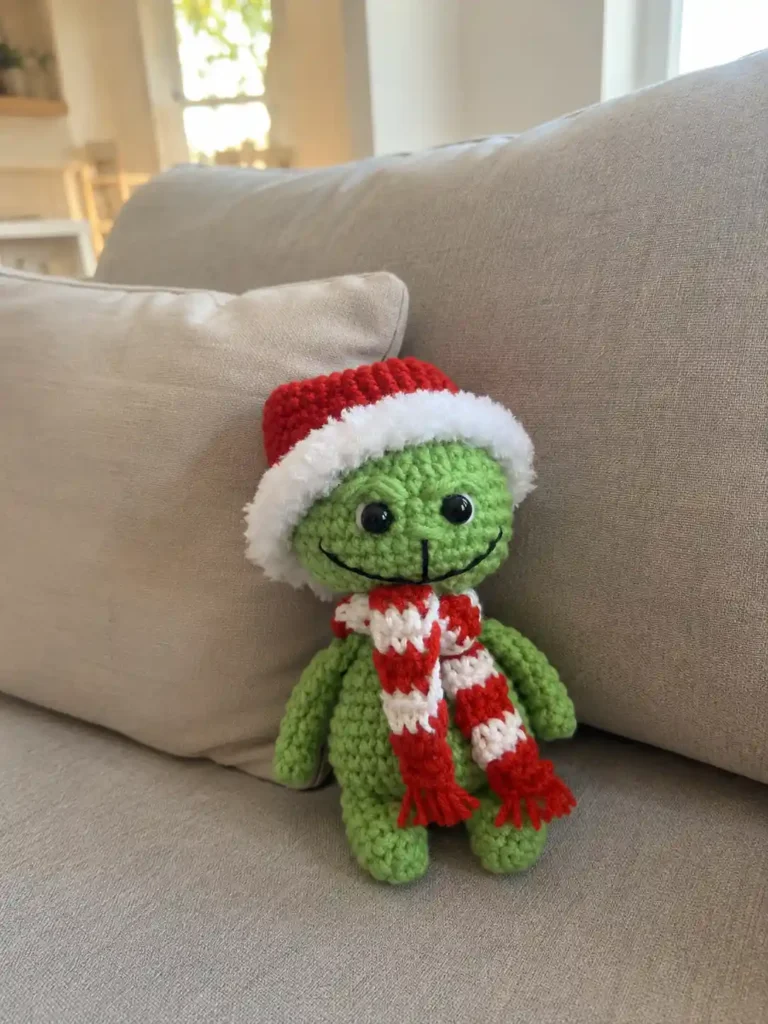 Green grinch amigurumi free crochet toy with red hat and striped scarf sitting on a beige couch cushion.