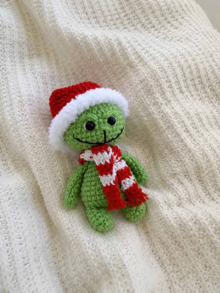 A completed green grinch amigurumi free crochet doll with a red Santa hat and scarf resting on a white knit blanket.