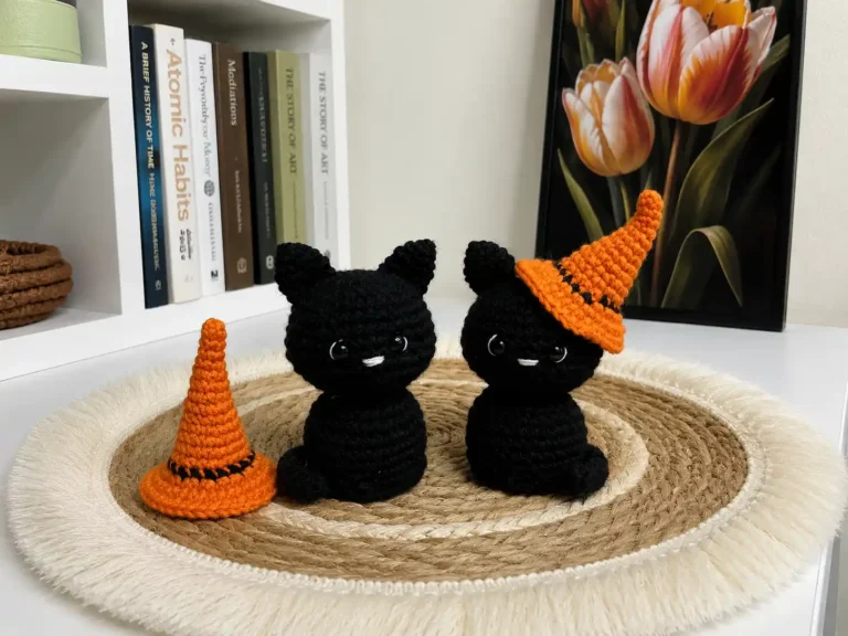 Two small black crochet cats on a wooden railing, one in a tiny orange witch hat, with a spare hat beside them.