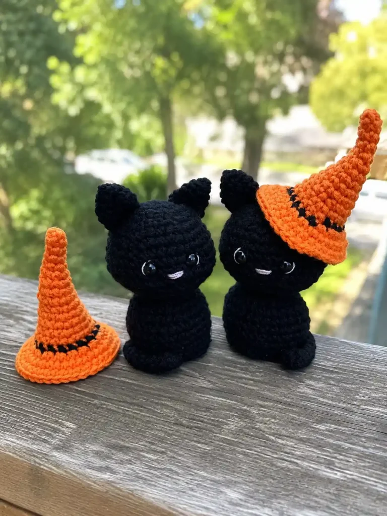 Two black yarn cats and an orange hat from a halloween black cat amigurumi pattern on a wooden patio with green trees.