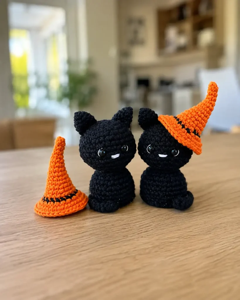Two black yarn cats and an orange hat from a halloween black cat amigurumi pattern, displayed standing on a wooden table.