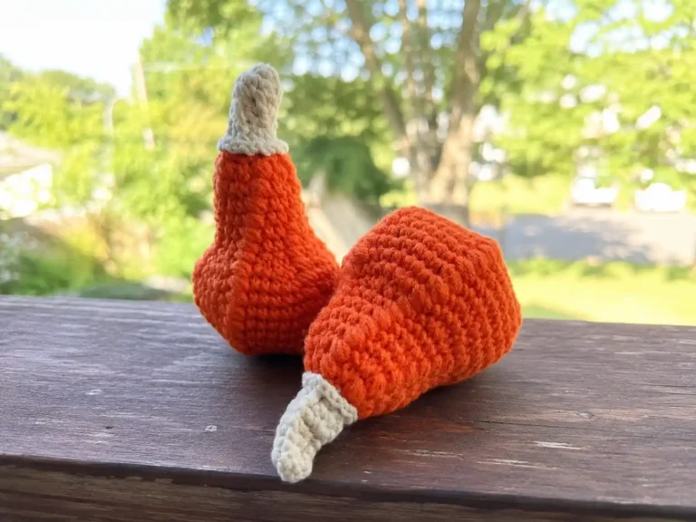 A hand in a cream sweater holds an orange crochet halloween pumpkin with a curly cream stem against greenery.