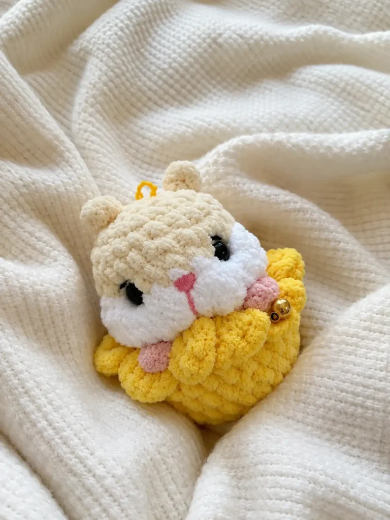 Plush cream and white yarn hamster in a flower amigurumi pattern resting on a soft white knit blanket with a gold bell.