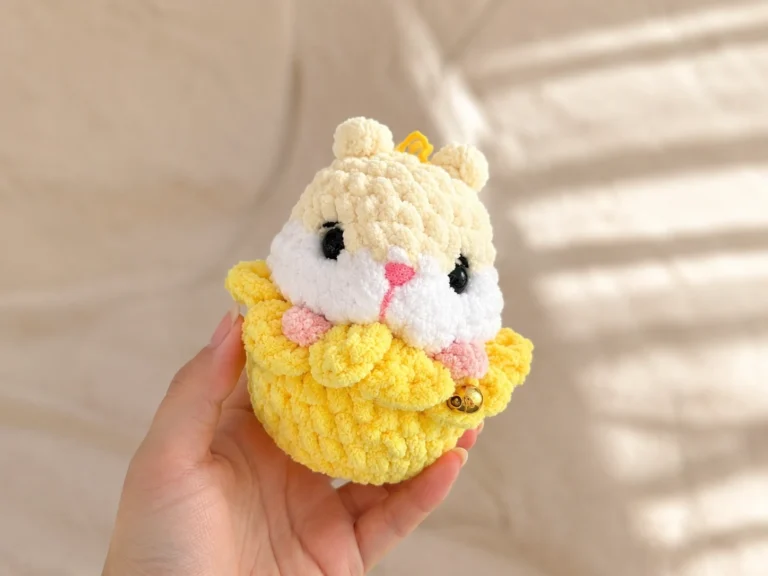 A cream and white crochet hamster amigurumi sitting inside a yellow crochet flower on a wooden railing outdoors.