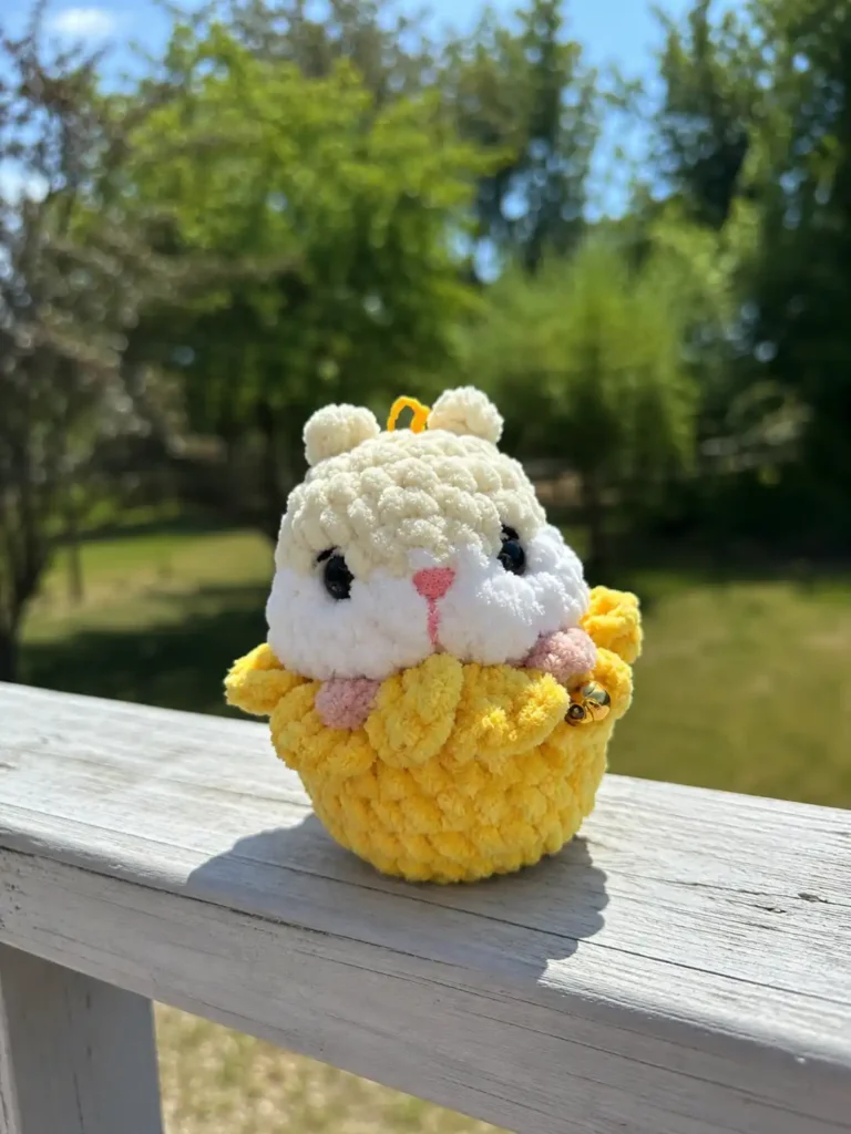 Plush chenille hamster in a flower amigurumi pattern with a bell sitting on a white outdoor railing near green trees.