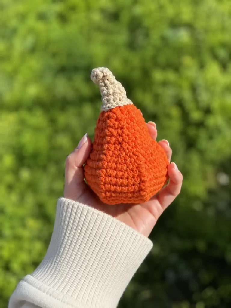 Hand in a white sleeve holding an orange gourd from a halloween pumpkin crochet pattern against a green backdrop.