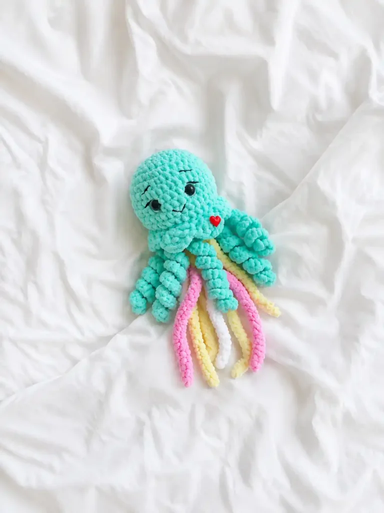 Top down view of a mint jellyfish amigurumi free pattern with pastel tentacles resting on a wrinkled white bedsheet.