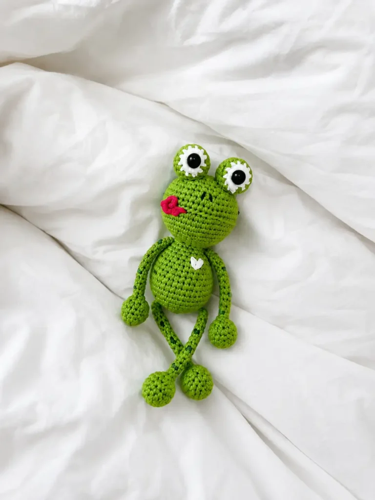 Green yarn kvaka the little froggy free crochet pattern lying flat on soft, wrinkled white bed sheets.