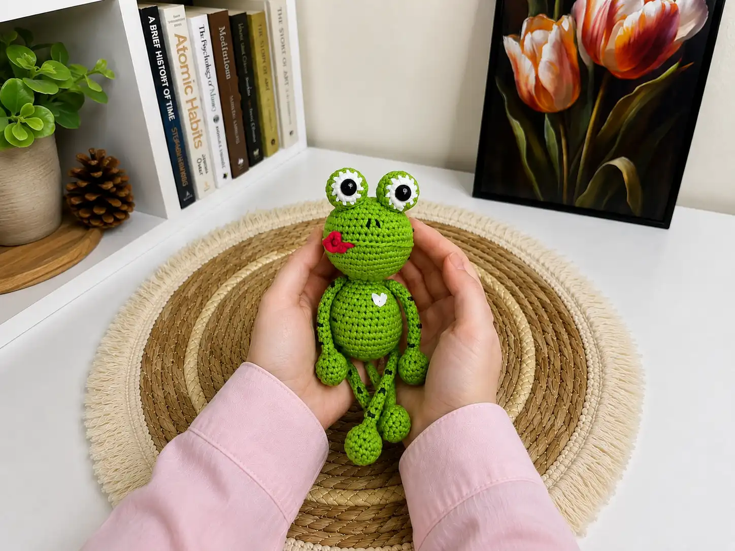 A hand holds a sage-green crochet frog amigurumi with white eyes and a red tongue against a grassy background.