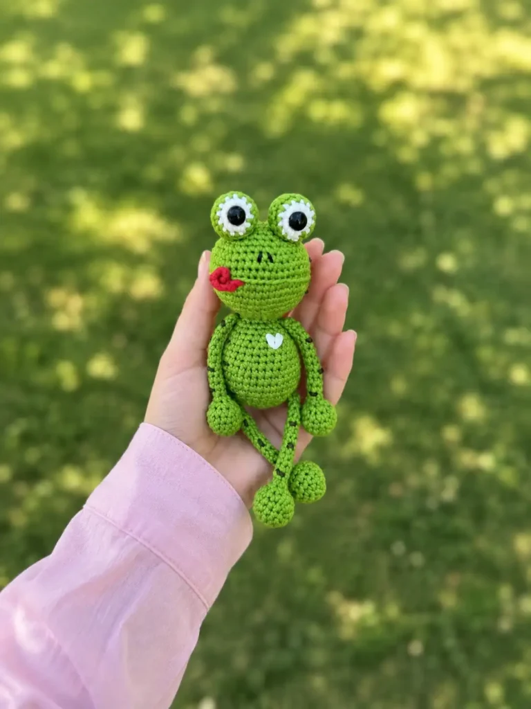Hand holding a green yarn kvaka the little froggy free crochet pattern outdoors against a blurred grass background.