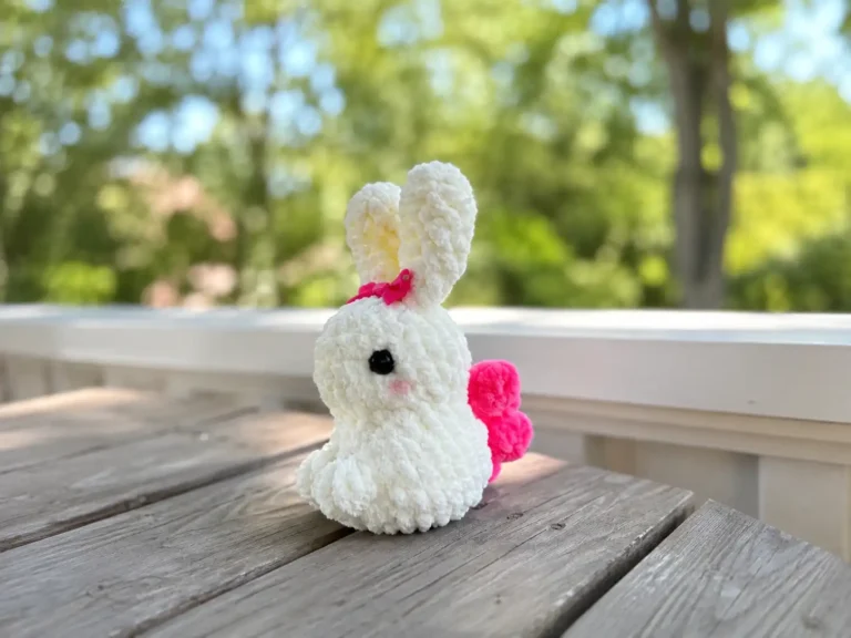 A cream chenille amigurumi bunny with black safety eyes, a pink bow, and bright pink wings, held in a hand.