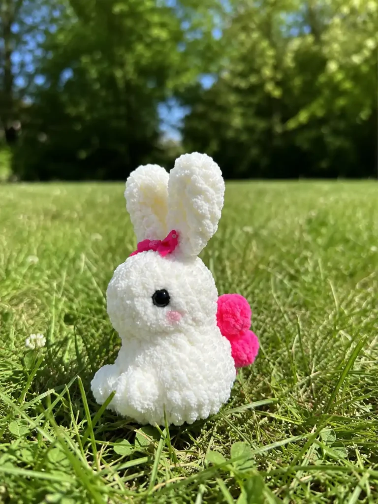 A white little bunny amigurumi free pattern sitting on green grass outdoors, showing its pink wings and textured yarn.
