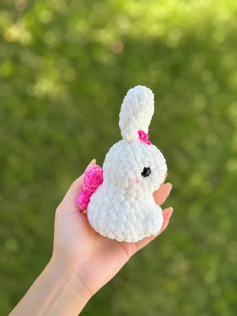 Hand holding the little bunny amigurumi free pattern with pink wings against a blurred green outdoor nature background.
