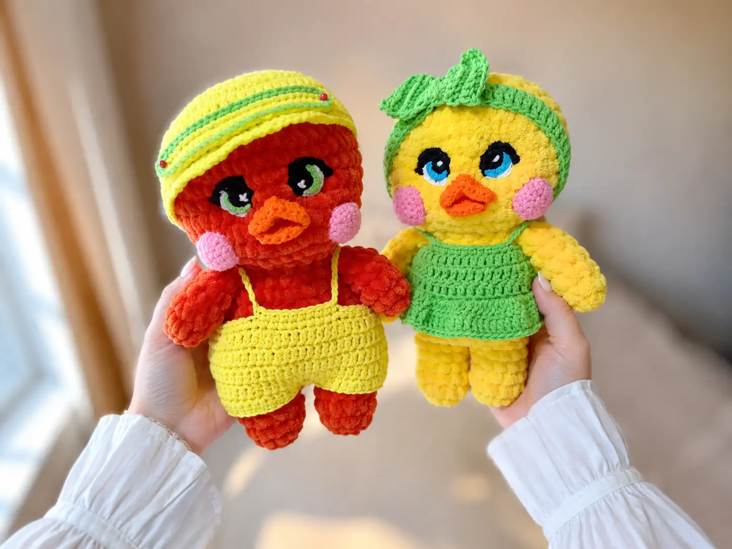 Lulu the yellow crochet duck in a green dress and headband beside Lolo the orange drake in overalls and a cap, by a plant.