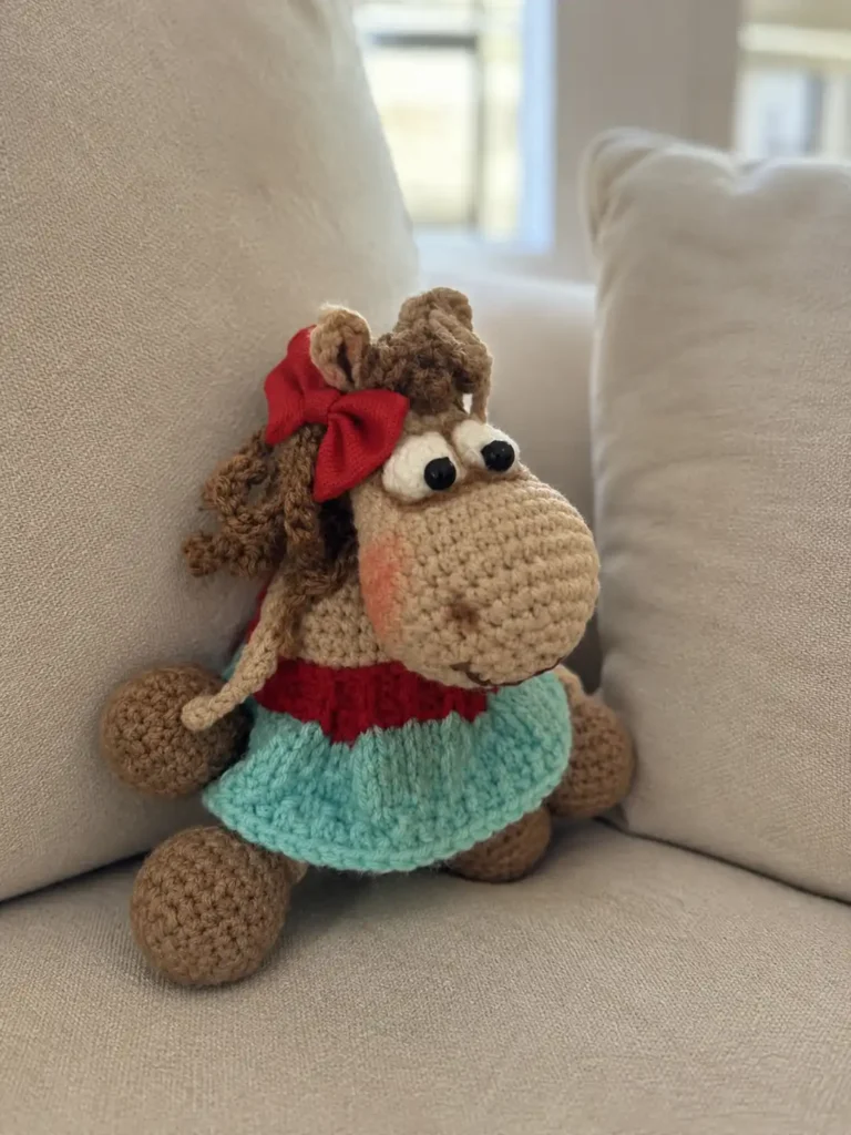 Marzipan sitting on a beige couch in a teal and red dress, crafted using this amigurumi horse free pattern.