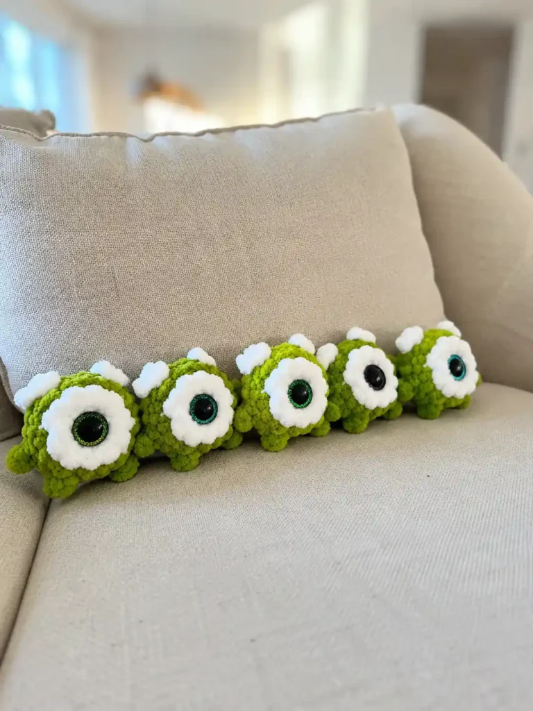 Five green plush one-eyed toys lined up on a beige couch, made using the mike wazowski amigurumi pattern free.