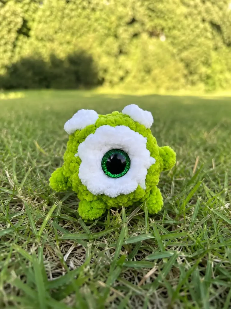 Green plush one-eyed toy sitting on bright green grass, crafted with the mike wazowski amigurumi pattern free.