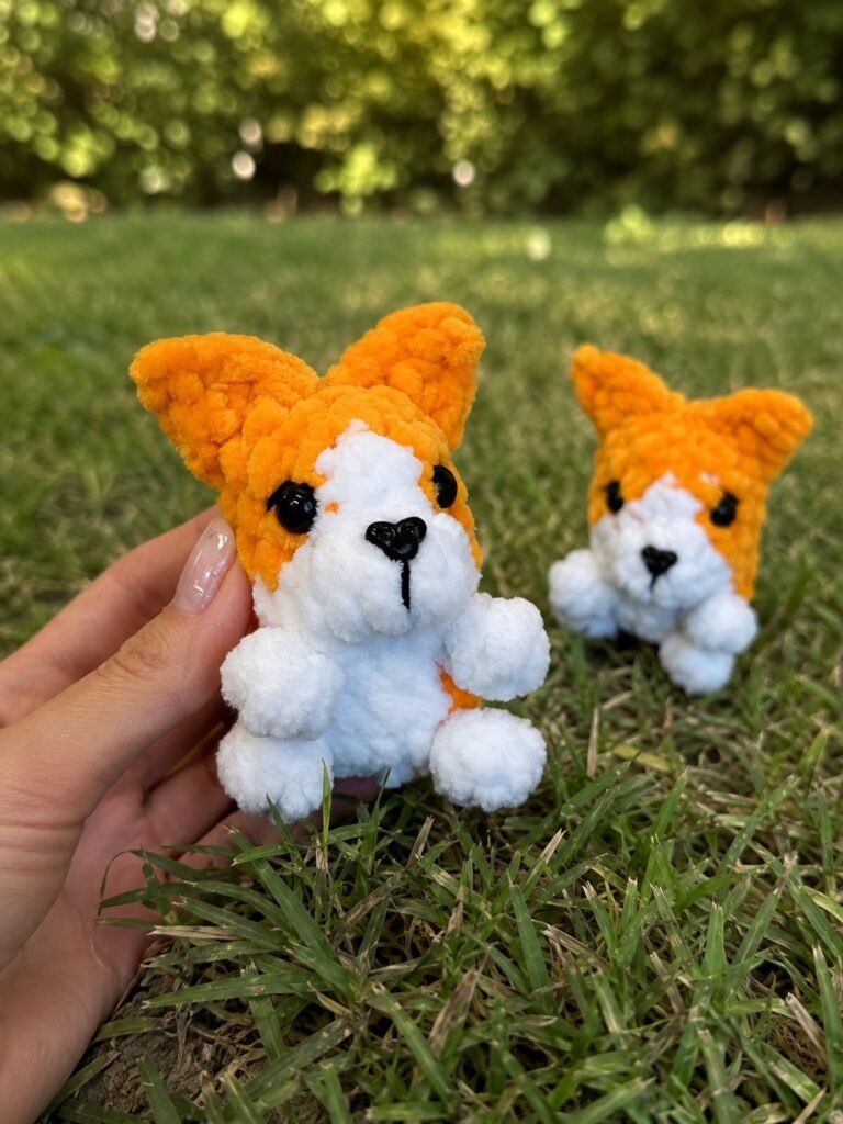 One hand holds an orange plush dog while another sits on green grass, showcasing the mini corgi amigurumi free pattern.