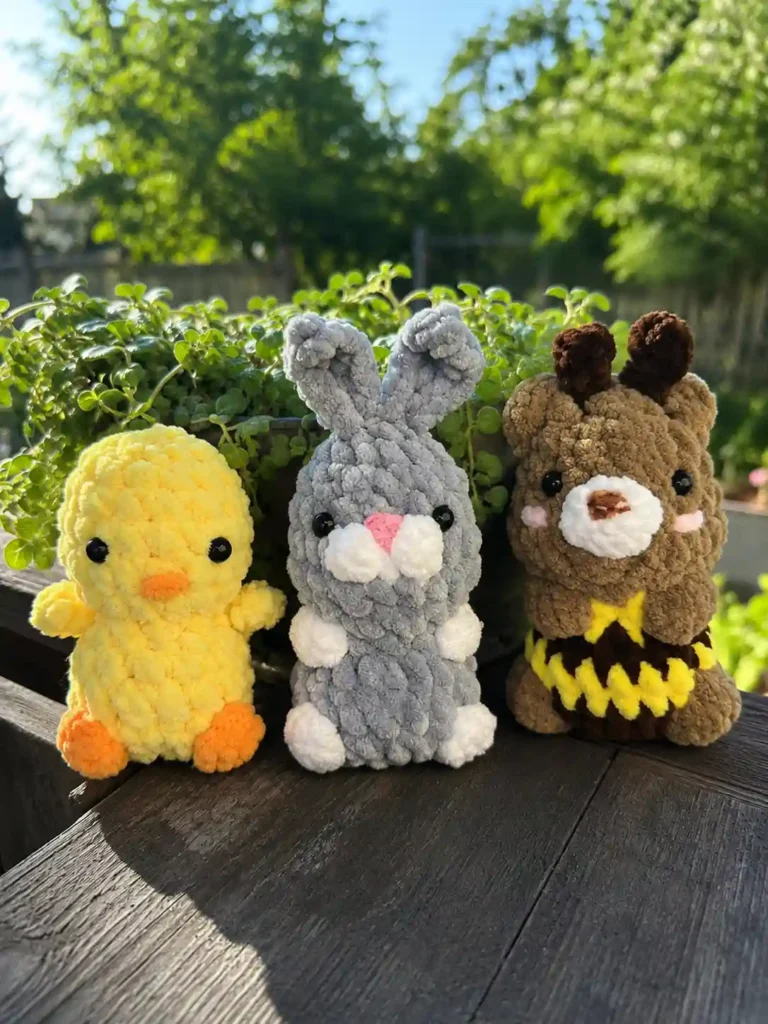 A plush yellow crochet duckling, a grey crochet bunny, and a brown crochet bear sitting side by side on a wooden ledge in the sunlight.