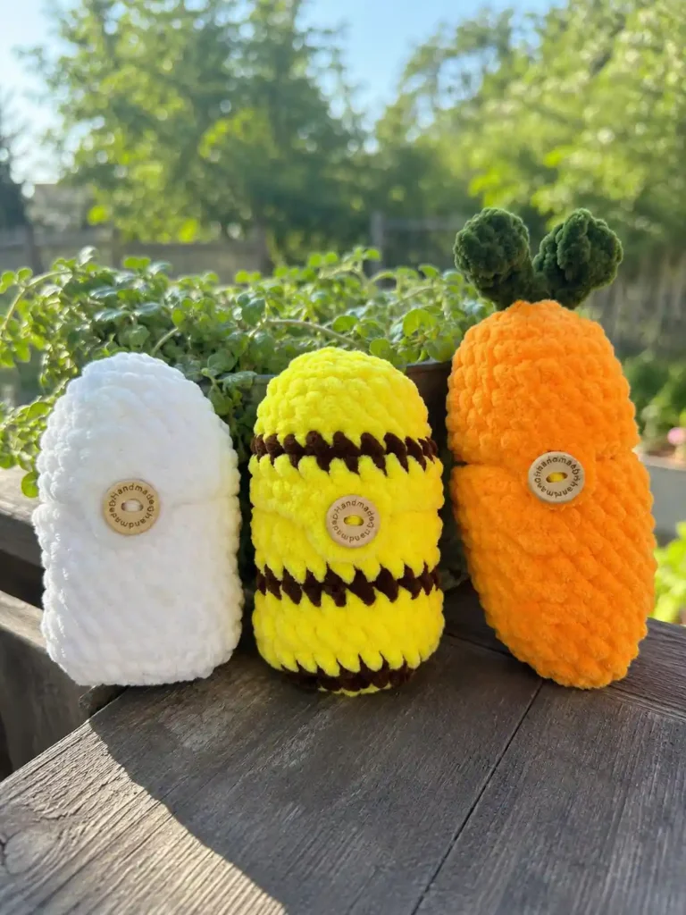 Three chunky closed crochet eggs—a white plain egg, a yellow and brown striped bee egg, and an orange carrot egg with green leaves—lined up on a wooden railing.