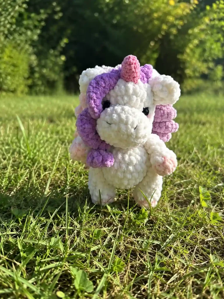 White chenille toy with purple mane from our mini unicorn crochet pattern free, standing on green grass in sunlight.