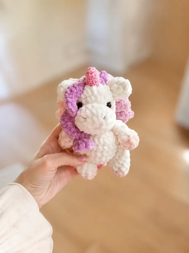 Hand holding a white plush toy with purple mane from our mini unicorn crochet pattern free, indoor background.