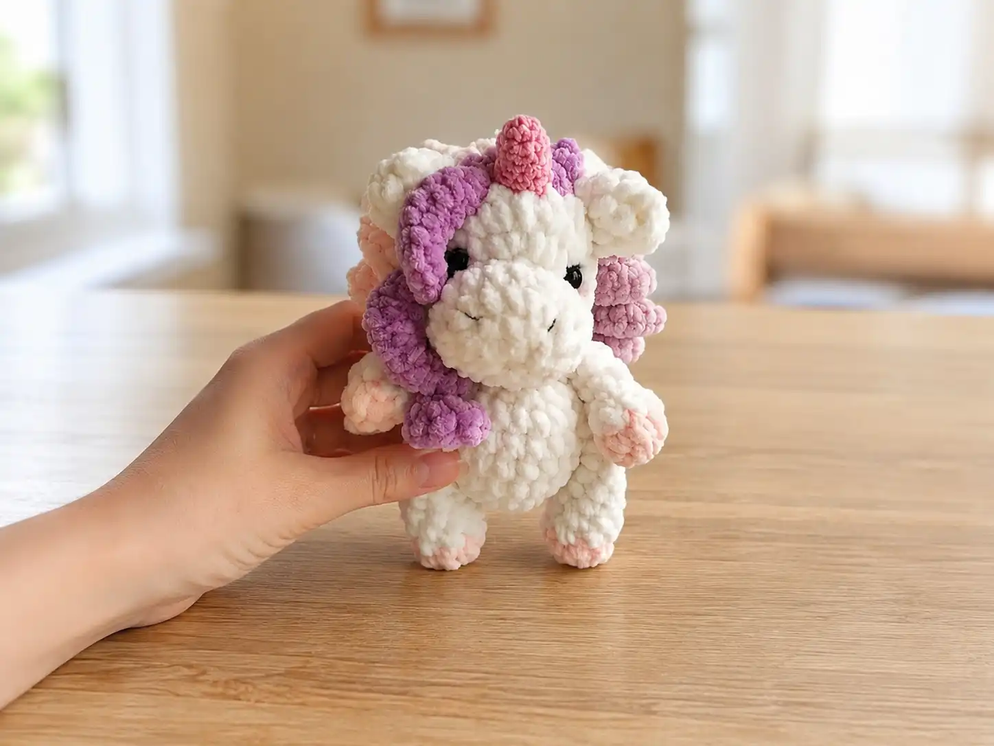A white crochet mini unicorn amigurumi with a purple mane and pink horn, held in a hand.