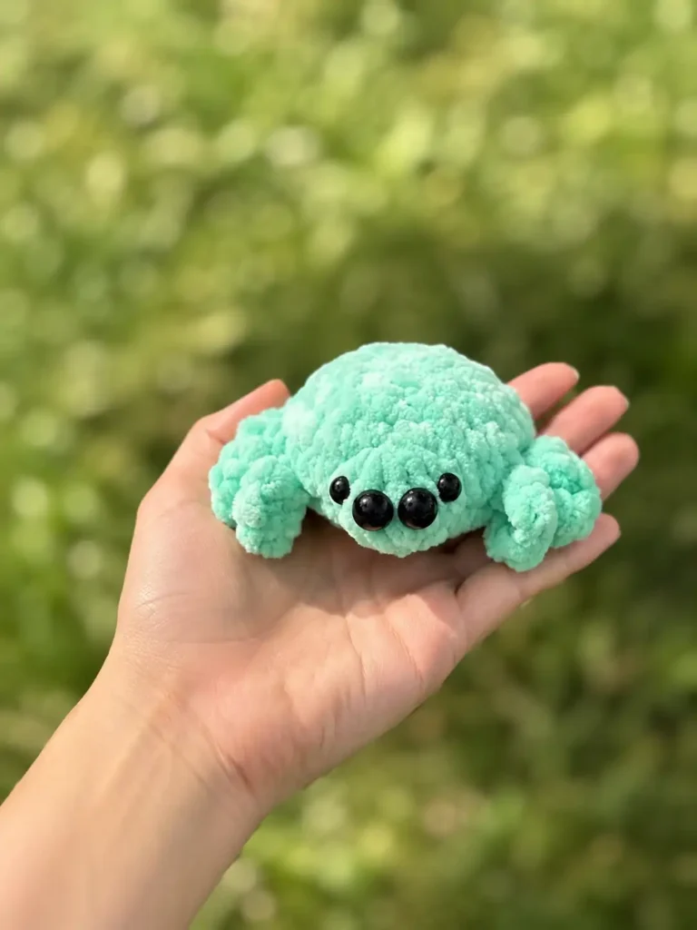 Mint green plush toy from a crochet spider amigurumi free pattern resting in an open palm over a green grass background.