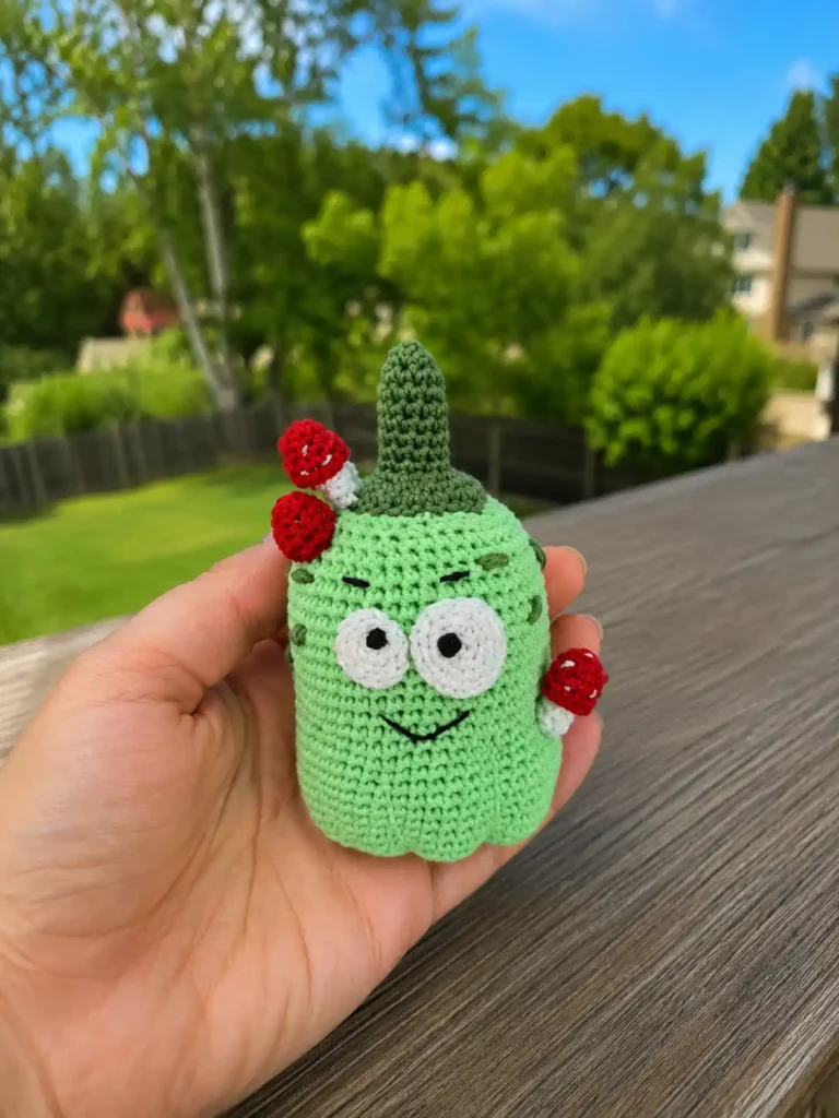 A hand holds a mint green yarn gourd with red mushrooms from the silly pumpkin amigurumi pattern outdoors.