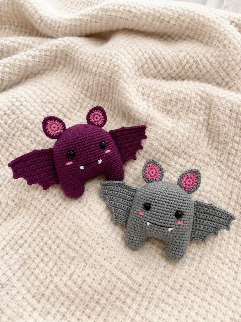 Two small crocheted bat amigurumi plushies in deep plum purple and soft gray with pink inner ears, black safety eyes, embroidered fangs, and open wings resting on a textured cream blanket.