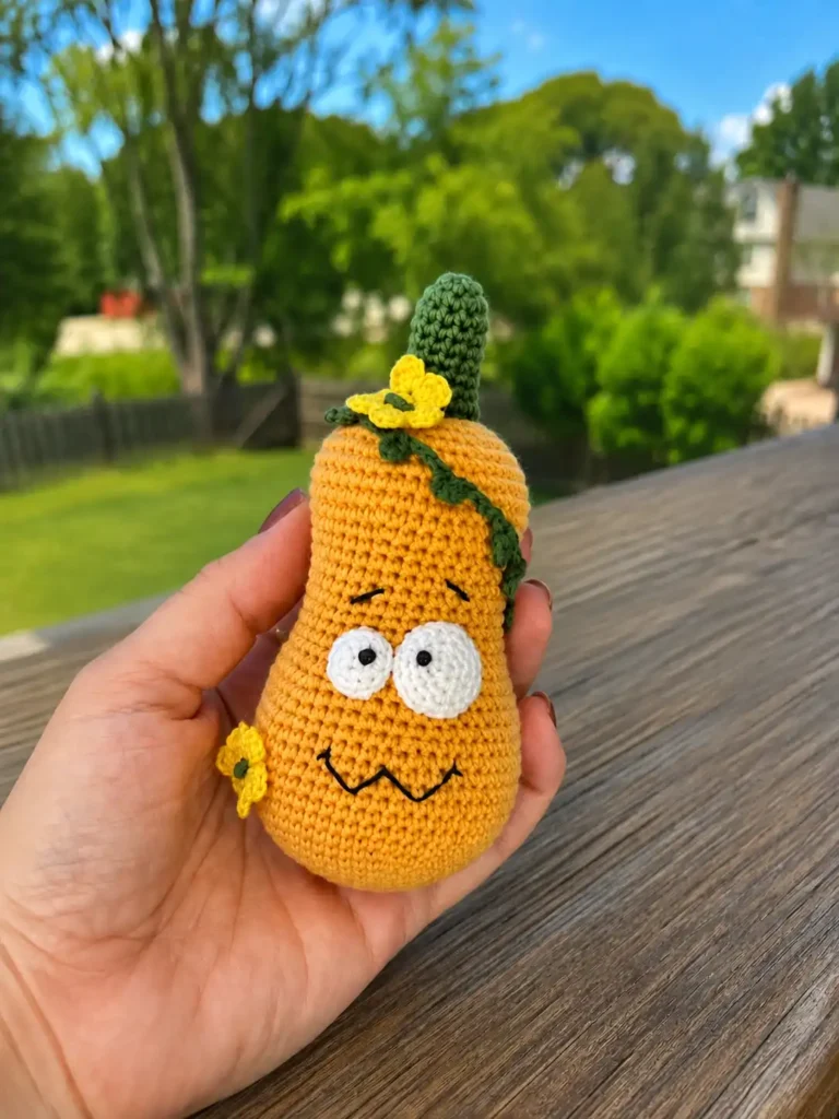 A hand displays a tall orange silly pumpkin amigurumi pattern featuring a green vine and yellow flowers outdoors on a deck.