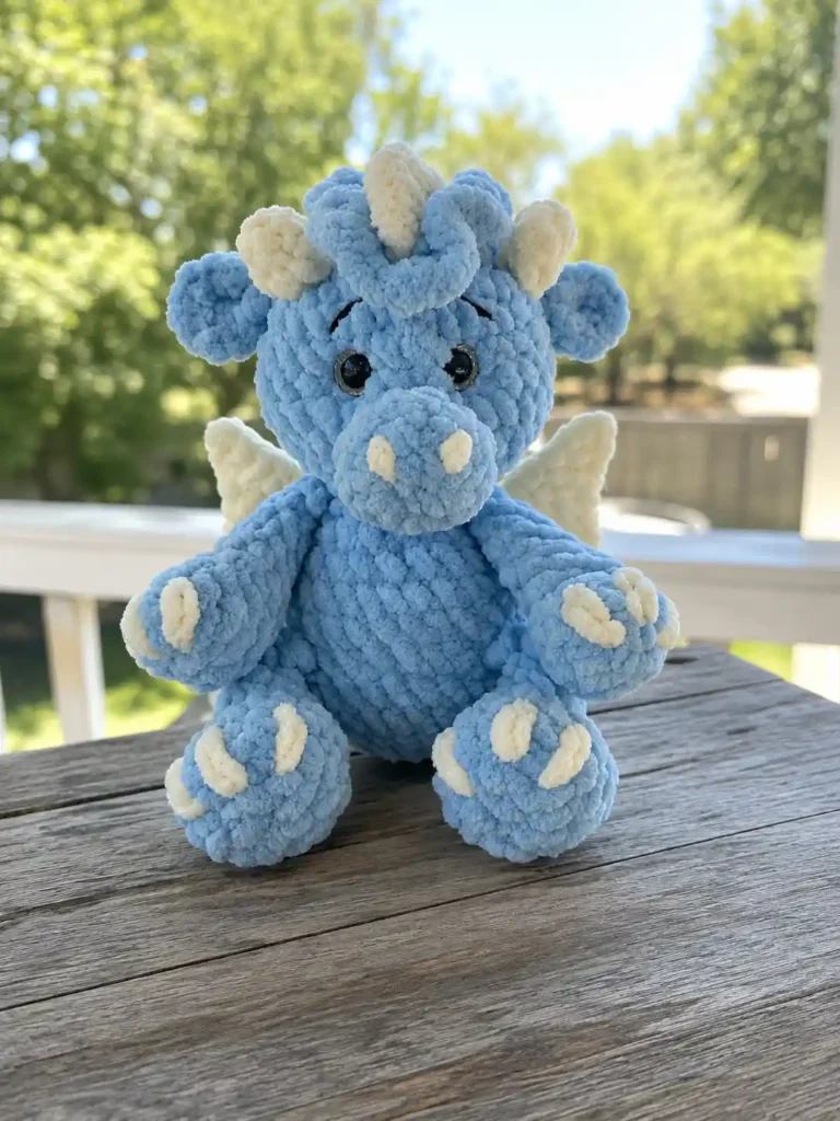 Blue chenille plush from a baby dragon crochet pattern free sitting on a slatted wooden outdoor table against green trees.