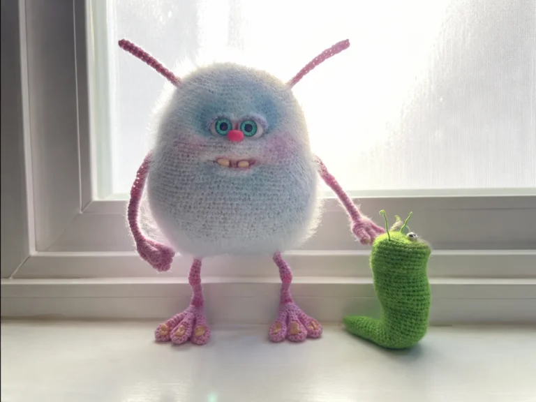 A white and pink crochet monster amigurumi with wire-posed arms and legs standing on a wood surface.