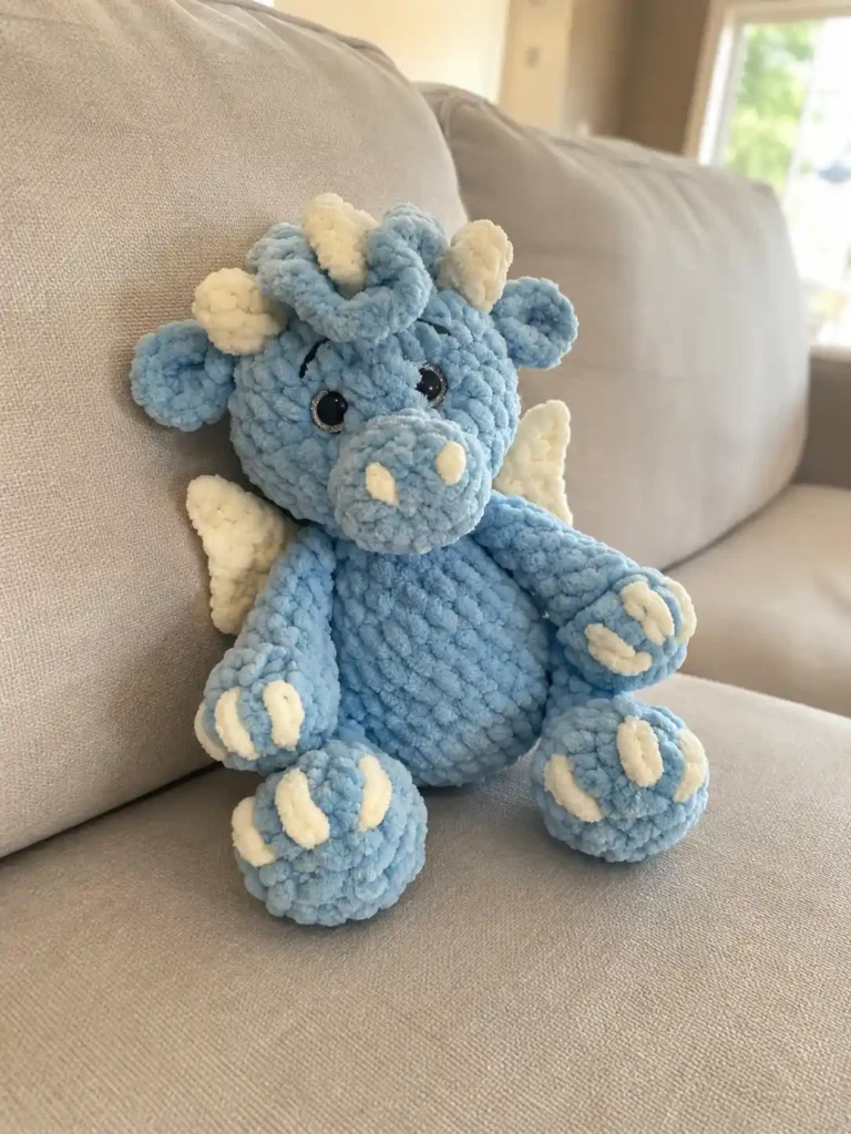 Blue yarn toy made from a baby dragon crochet pattern free resting on a beige textured couch with cream wings visible.