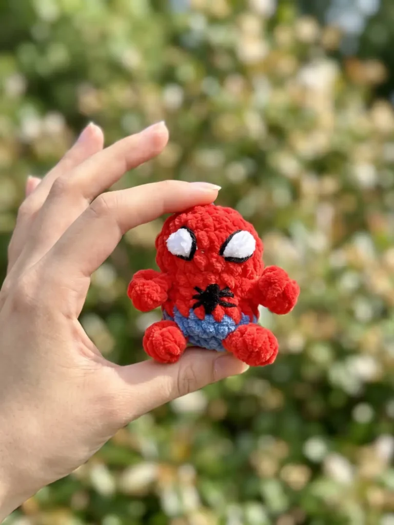 Hand holding a red and blue chenille toy from a spiderman amigurumi keychain free pattern against blurred green leaves.