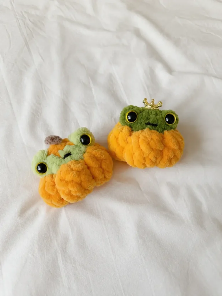 Two squishy frog in a pumpkin amigurumi pattern toys, one with a crown and one with a stem, resting on a white sheet.