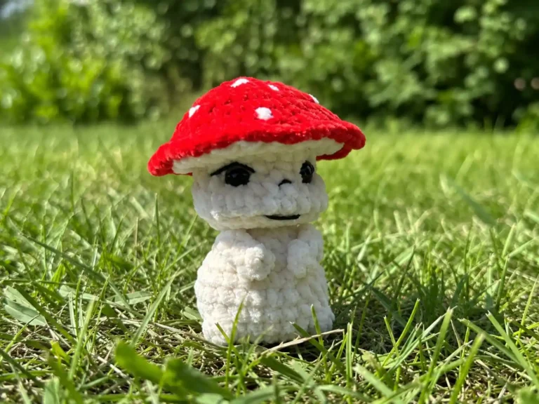 A crochet toadstool mushroom amigurumi with a red spotted cap, shown in green grass and on a wooden table.