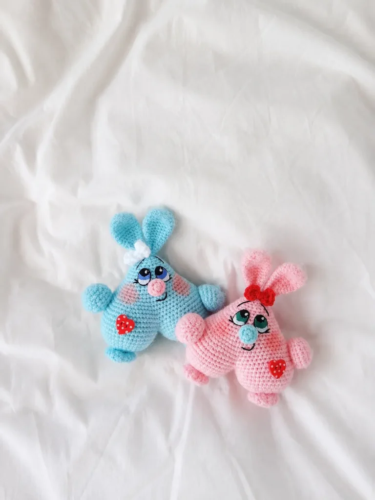 Blue and pink yarn valentine heart bunnies amigurumi pattern free, displayed flat on a wrinkled white sheet background.
